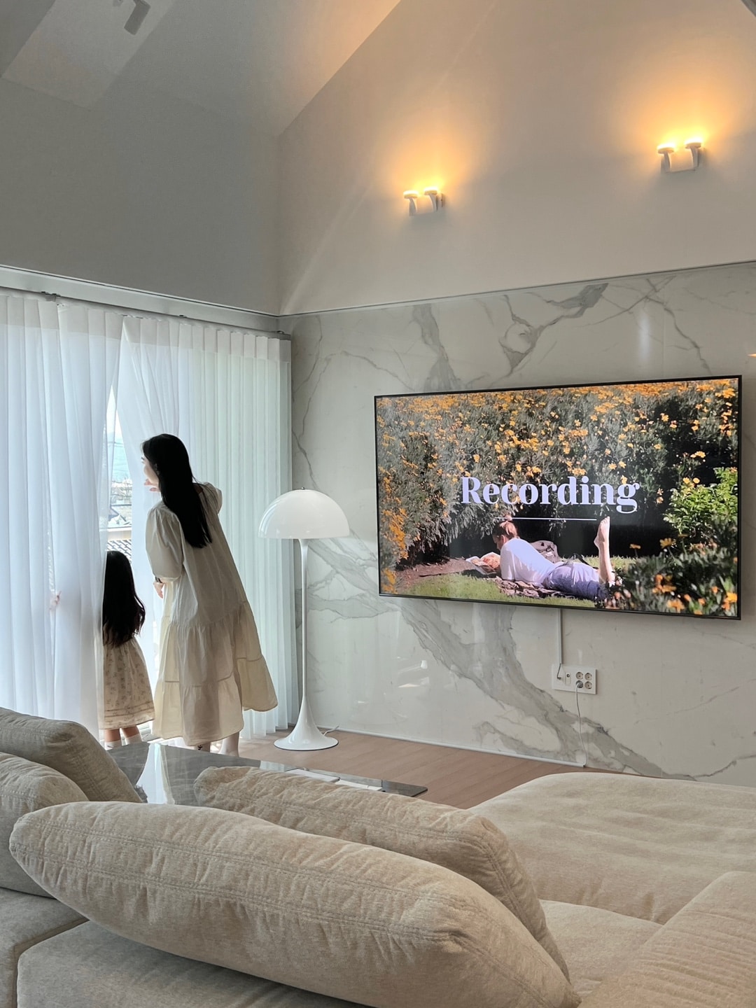 A living room is displayed with a modern design, featuring a large screen mounted on the wall that reads 'Recording.' A woman and a child stand by sheer drapes, gazing outside. A soft sofa and a contemporary lamp complete the inviting space.