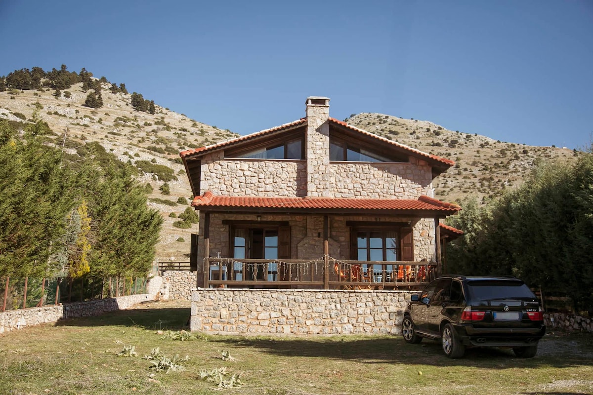 A charming stone house is set against a backdrop of rolling hills, featuring a sloped roof and large windows. A spacious porch with railings extends in front, while a parked SUV is visible in the foreground, indicating accessible parking.