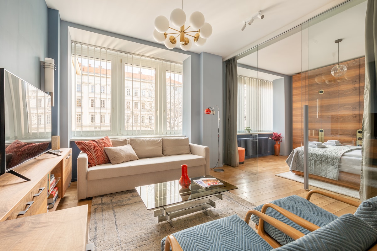A modern living area displays a light-colored sofa with decorative cushions, accompanied by a glass coffee table. Soft textures are introduced by a woven rug and a cozy chair. Large windows allow natural light to fill the space, with a view of the street visible outside.