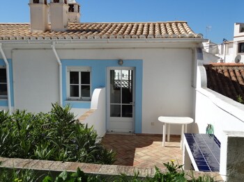 The exterior showcases a charming cottage entrance, featuring a white façade with blue accents. A small table is positioned on a tiled terrace surrounded by greenery. The sunny sky highlights the cottage's inviting ambiance.