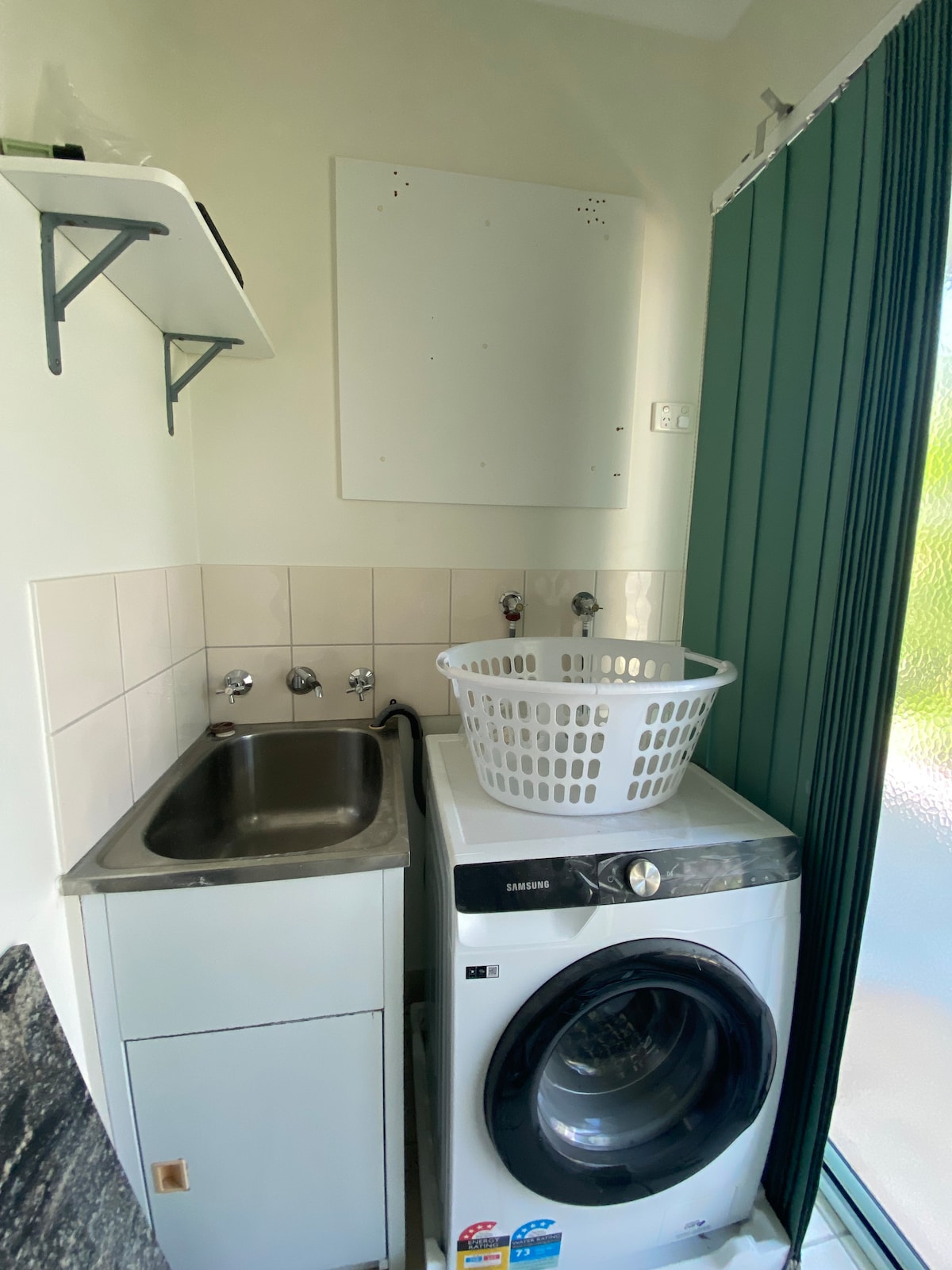 A compact laundry area is presented, featuring a stainless steel sink adjacent to a washing machine. A white laundry basket is placed on top of the machine. Shelves above provide storage, and a green curtain partially covers a window, allowing natural light to enter.