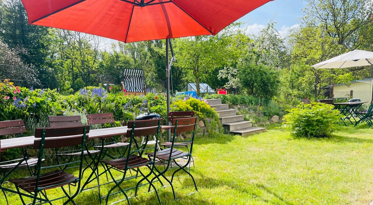 A spacious garden area is shown, featuring multiple wooden tables and chairs shaded by large red umbrellas. Vibrant flowers and greenery surround the garden, creating a serene outdoor space. Stone steps lead to a higher level, enhancing the inviting atmosphere of the setting.