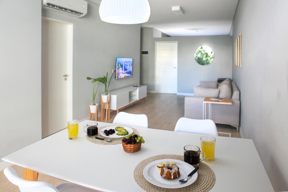 A modern dining area is shown, equipped with a white table set for breakfast, featuring glasses of juice and a bowl of fruit. In the background, a comfortable living room is visible, complete with a sofa and a wall-mounted television.