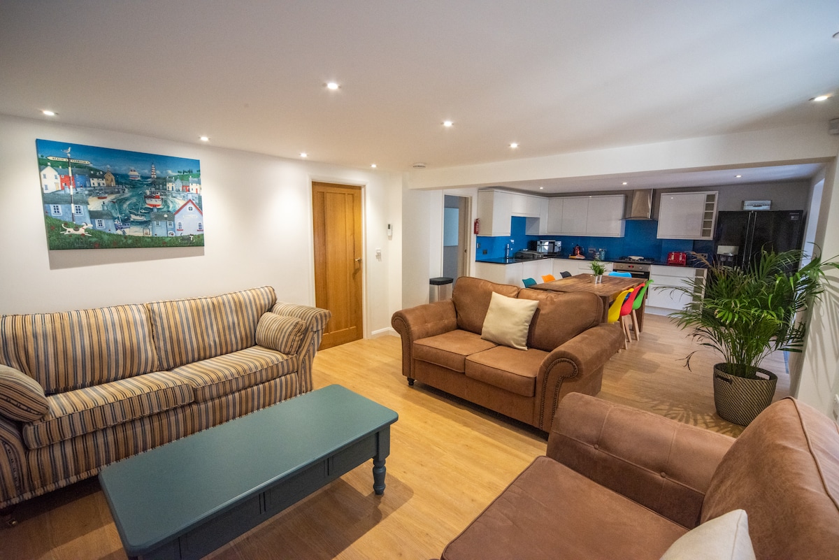 The open-plan living area features three comfortable sofas arranged around a coffee table. A colorful painting, depicting a coastal scene, adorns the wall. The modern kitchen with blue accents and integrated appliances is visible in the background, enhancing the light-filled space.