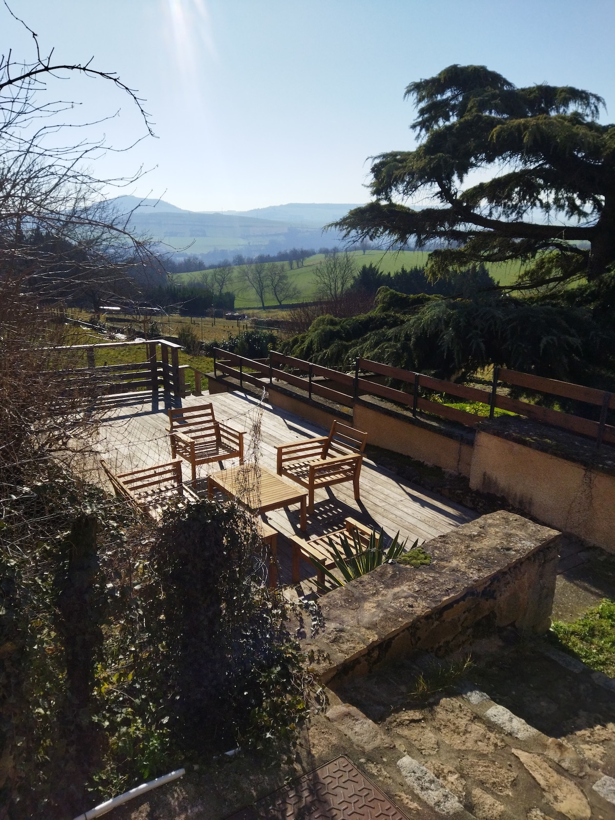 A wooden terrace is visible, featuring four chairs arranged around a small table. Lush greenery frames the space, with expansive views of the rolling hills in the background. The setting captures a serene outdoor atmosphere, ideal for relaxation.