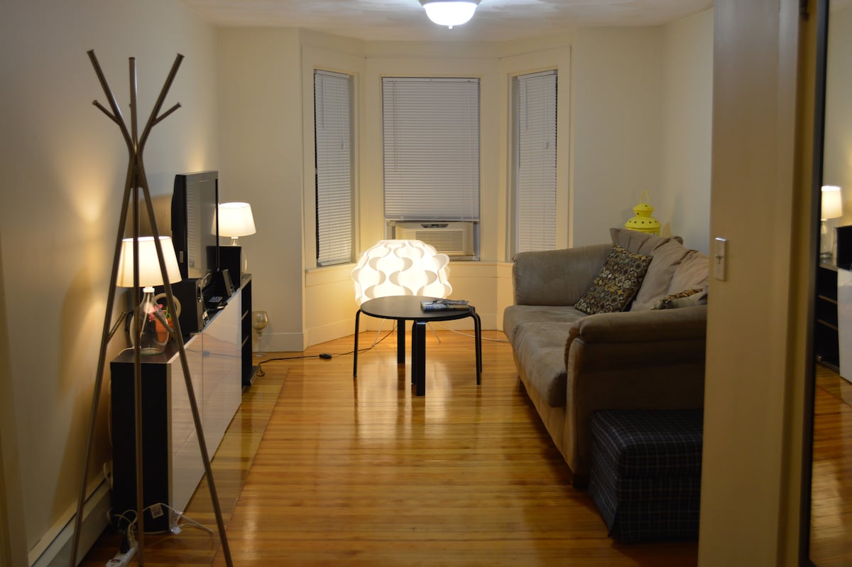 A living area featuring natural wood flooring is illuminated by lamps. A couch is positioned near the center alongside a round table. Large windows allow natural light, complemented by elegant curtains. A TV stand with a flat-screen television is visible, adding convenience to the space.