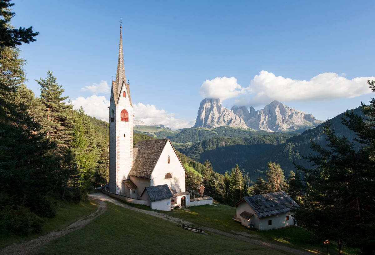 A charming church with a tall steeple is nestled among green trees, framed by majestic mountains in the background. Soft clouds hover above, adding depth to the serene landscape, which is accessible via a winding pathway leading towards the structure.
