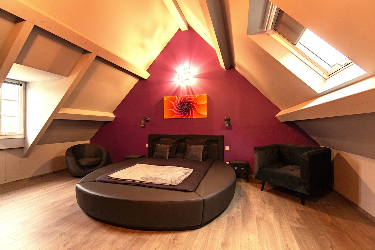 An elegant attic room features a spacious circular bed, surrounded by sloped ceilings and wooden beams. A vivid artwork adorns the wall, while soft lighting enhances the ambiance. Two plush armchairs are positioned nearby, providing a comfortable seating area. Natural light floods in through a skylight.