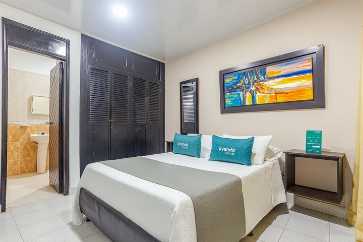 A spacious bedroom features a large bed covered with a soft white blanket and grey throw. A vibrant piece of artwork hangs above, contrasting against the dark wood furnishings, including a nightstand. A bathroom door is visible in the background, complementing the room's layout.