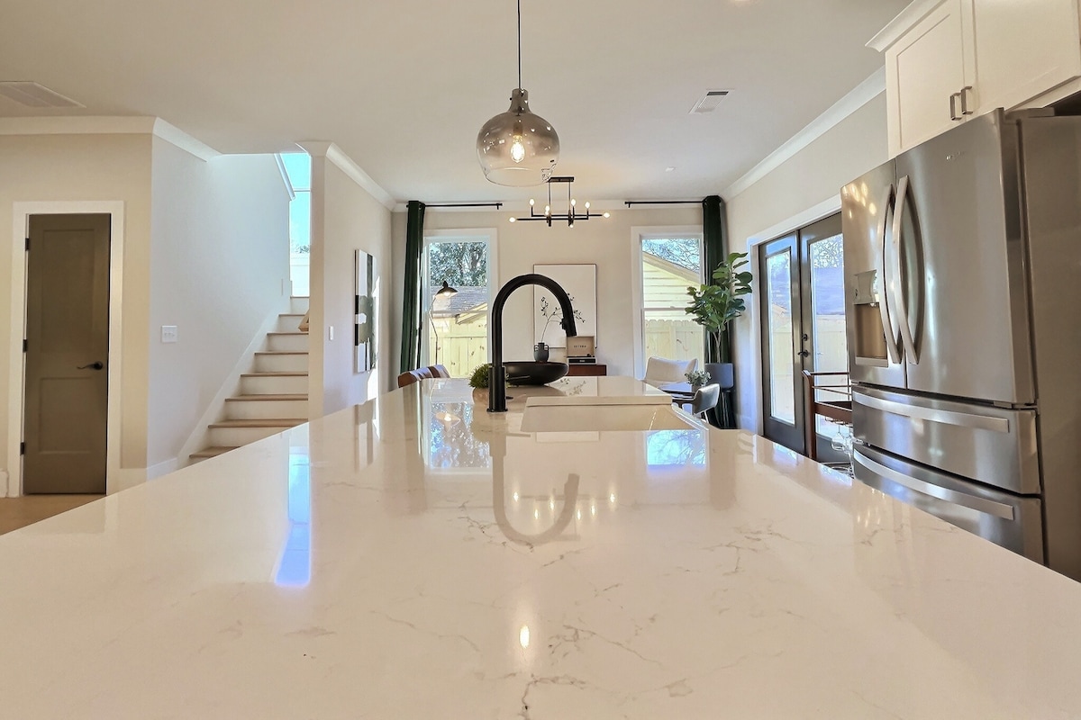 A modern kitchen features a light marble countertop, complemented by sleek stainless-steel appliances and a stylish faucet. Natural light fills the space through large glass doors, connecting to an outdoor area. In the background, a staircase leads to the upper level, enhancing the open layout.