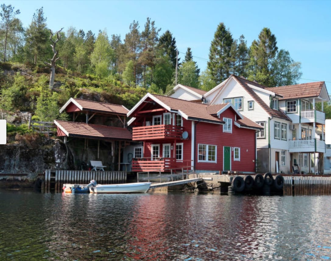 A collection of lakeside structures is depicted, featuring a red wooden building with a sloped roof and several windows. A smaller covered area is visible beside the main building, with a small white boat docked at the jetty. Lush greenery surrounds the site, enhancing the natural setting.