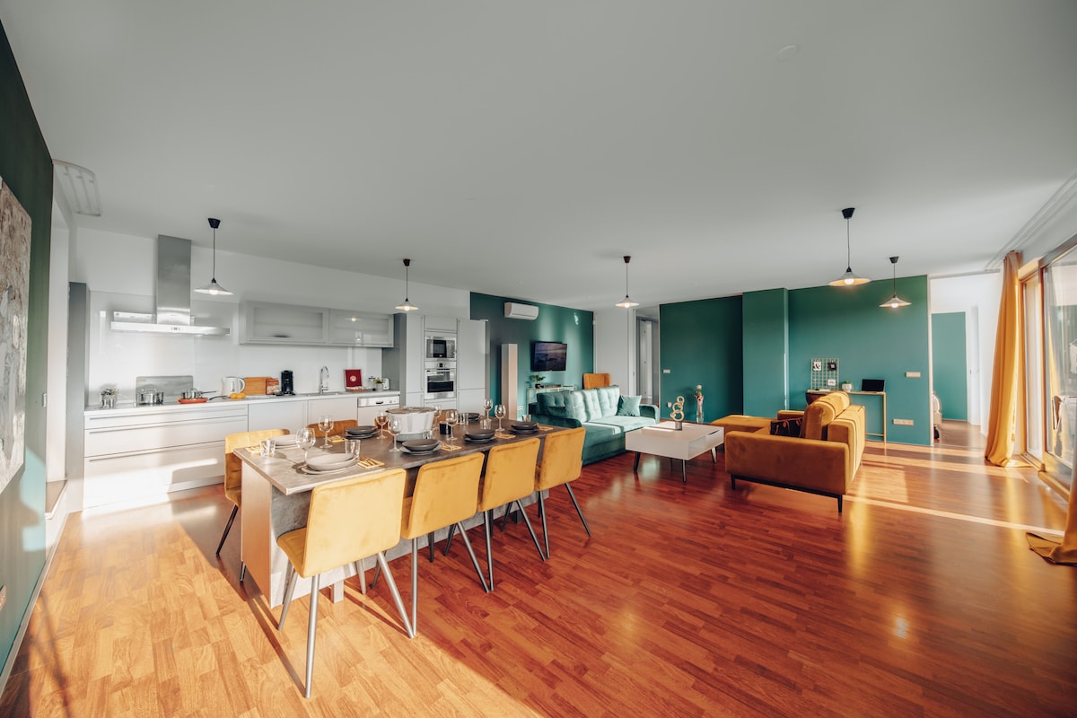 An open-plan living area showcases a dining table surrounded by eight yellow chairs and a well-appointed kitchen in the background. Two sofas in soft tones and a large window allow natural light to fill the space, highlighting the warm wooden flooring.