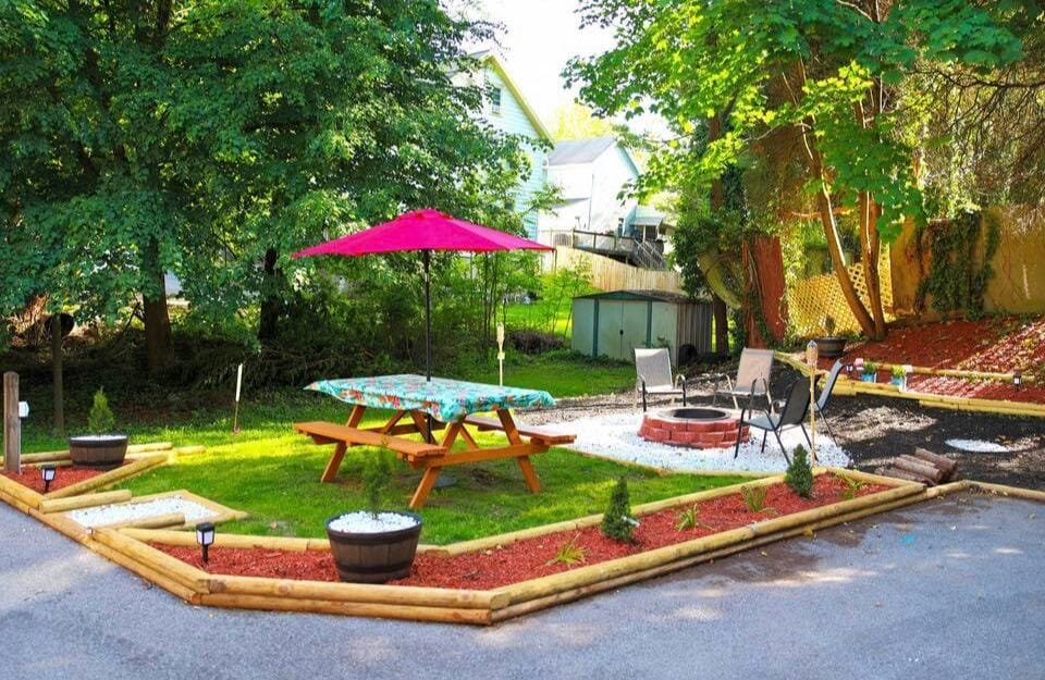 An outdoor space is featured with a picnic table shaded by a pink umbrella and surrounded by landscaped borders. A fire pit with seating is visible nearby, nestled among greenery. Soft lighting fixtures enhance the area, creating a welcoming environment for outdoor gatherings.