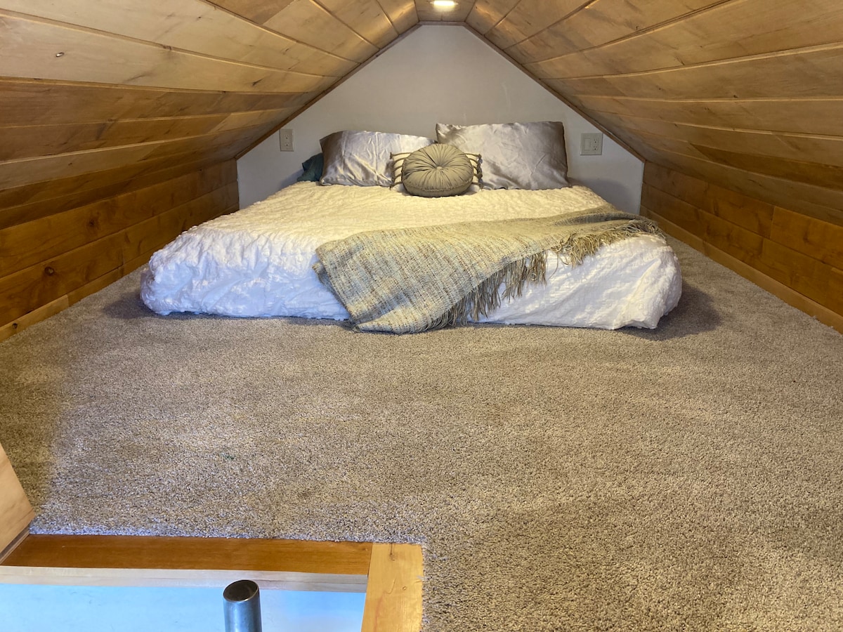 A cozy loft space features a large sleeping area with a plush carpeted floor and a spacious bed adorned with light bedding and decorative pillows. Exposed wooden beams create a warm, rustic ambiance, while a soft throw blanket is draped across the bed.