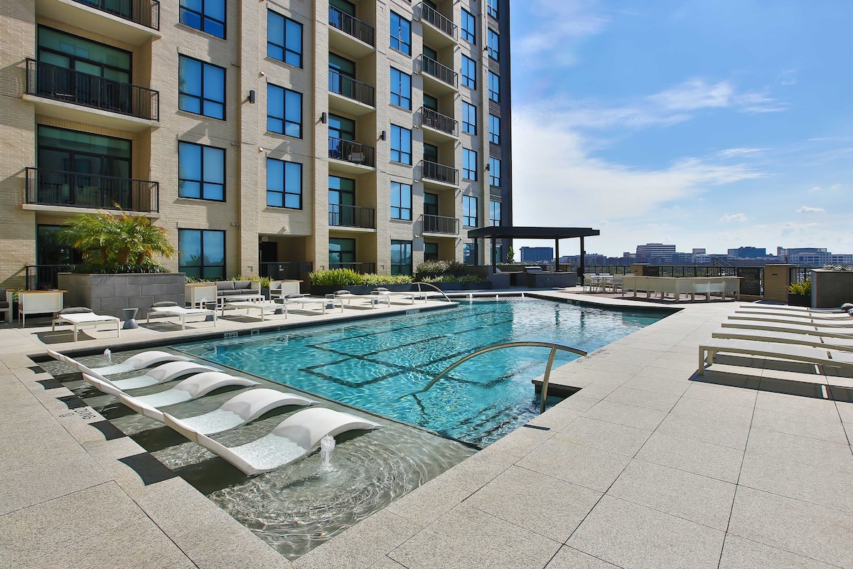An outdoor swimming pool is framed by modern lounge chairs and a shaded seating area. The pool features a relaxing waterfall effect, with clear blue water reflecting the bright sky. The surrounding concrete patio is designed for leisure and comfort.