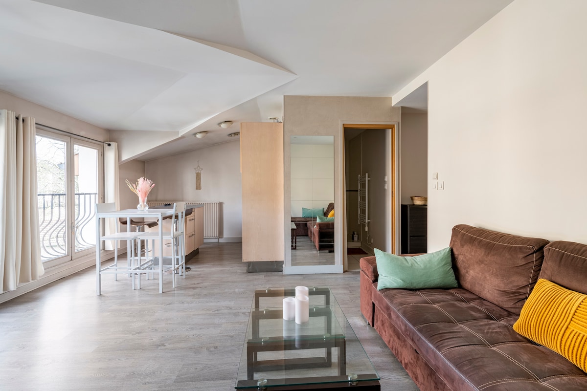 A spacious living area features a comfortable brown sofa with colorful cushions. A coffee table sits in front of the sofa, and a dining table with high chairs is visible in the background. Large windows allow natural light to fill the room.