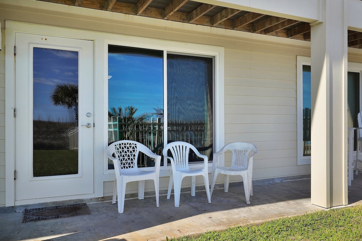 A private outdoor area features three white chairs arranged on a concrete patio, with sliding glass doors leading inside. The backdrop showcases a well-maintained grassy area and a glimpse of palm trees against a clear blue sky.