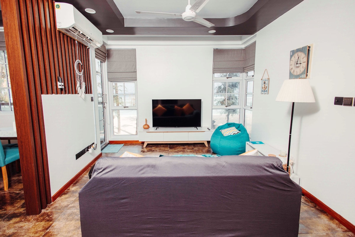 A living area features a comfortable dark sofa facing a wall-mounted television. Natural light floods the space through multiple windows with light curtains. A ceiling fan hangs above, and a stylish lamp stands next to a vibrant blue bean bag, adding a touch of color.