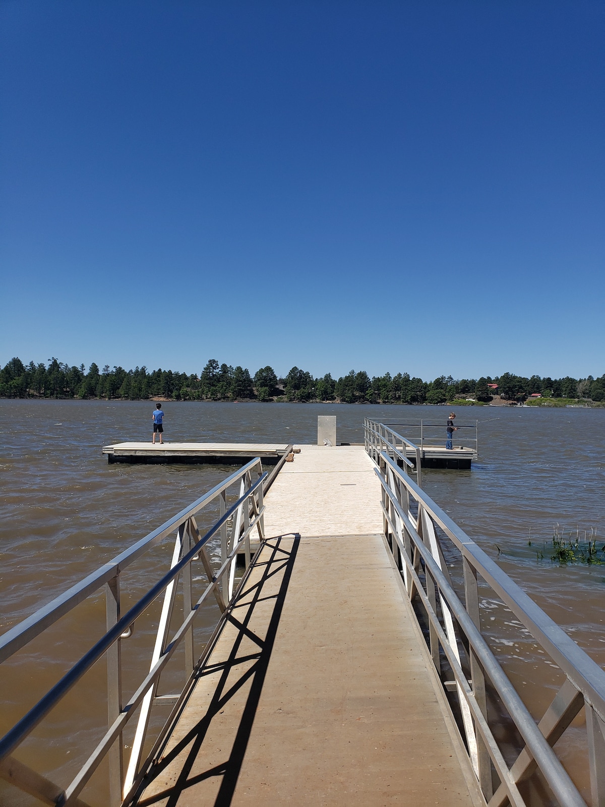 A wooden dock extends into the calm waters of a lake, flanked by metal railings. Two figures stand on separate platforms at its end, surrounded by a backdrop of trees and clear blue skies. The serene setting offers a sense of tranquility.