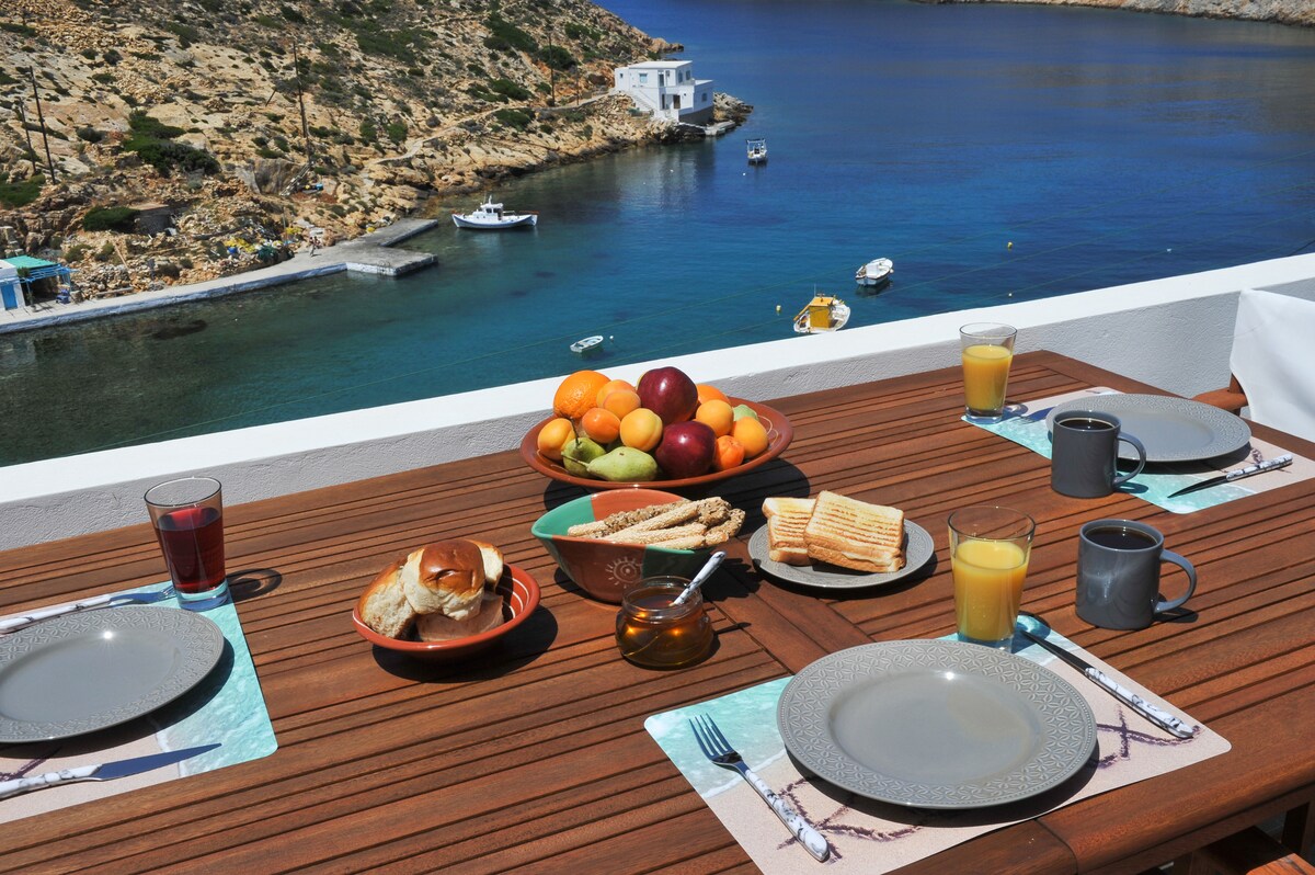 A dining table is set with plates and cups, overlooking tranquil waters. Fresh fruits are displayed in a bowl, accompanied by breakfast items including sandwiches and pastries. The serene view includes boats anchored nearby and the coastline in the background.