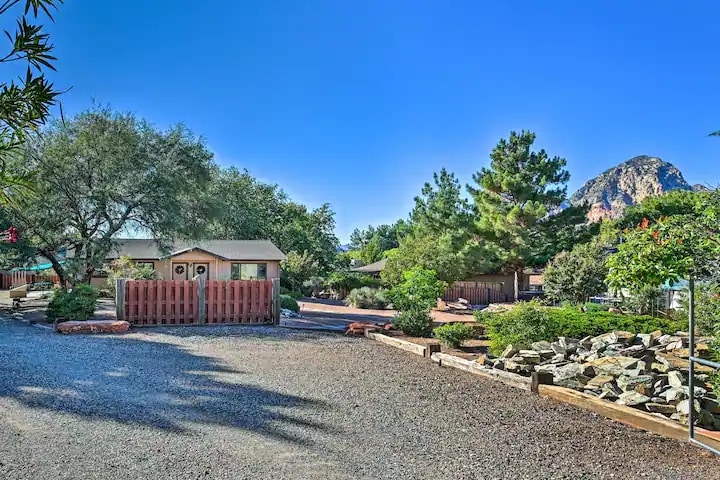 A gravel driveway leads to a charming duplex set against a clear blue sky. Lush trees and shrubs frame the property, offering a sense of tranquility. A vibrant red fence borders the front yard, while distant rock formations provide an appealing natural backdrop.