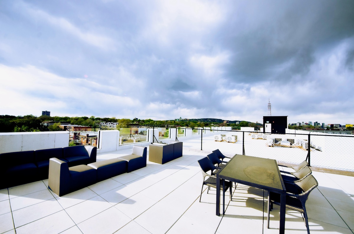 The rooftop terrace is equipped with comfortable seating arrangements, featuring a dark lounge set and a dining table. The space is surrounded by a safety railing, offering expansive views of the surrounding area, complemented by a cloudy sky and distant greenery.