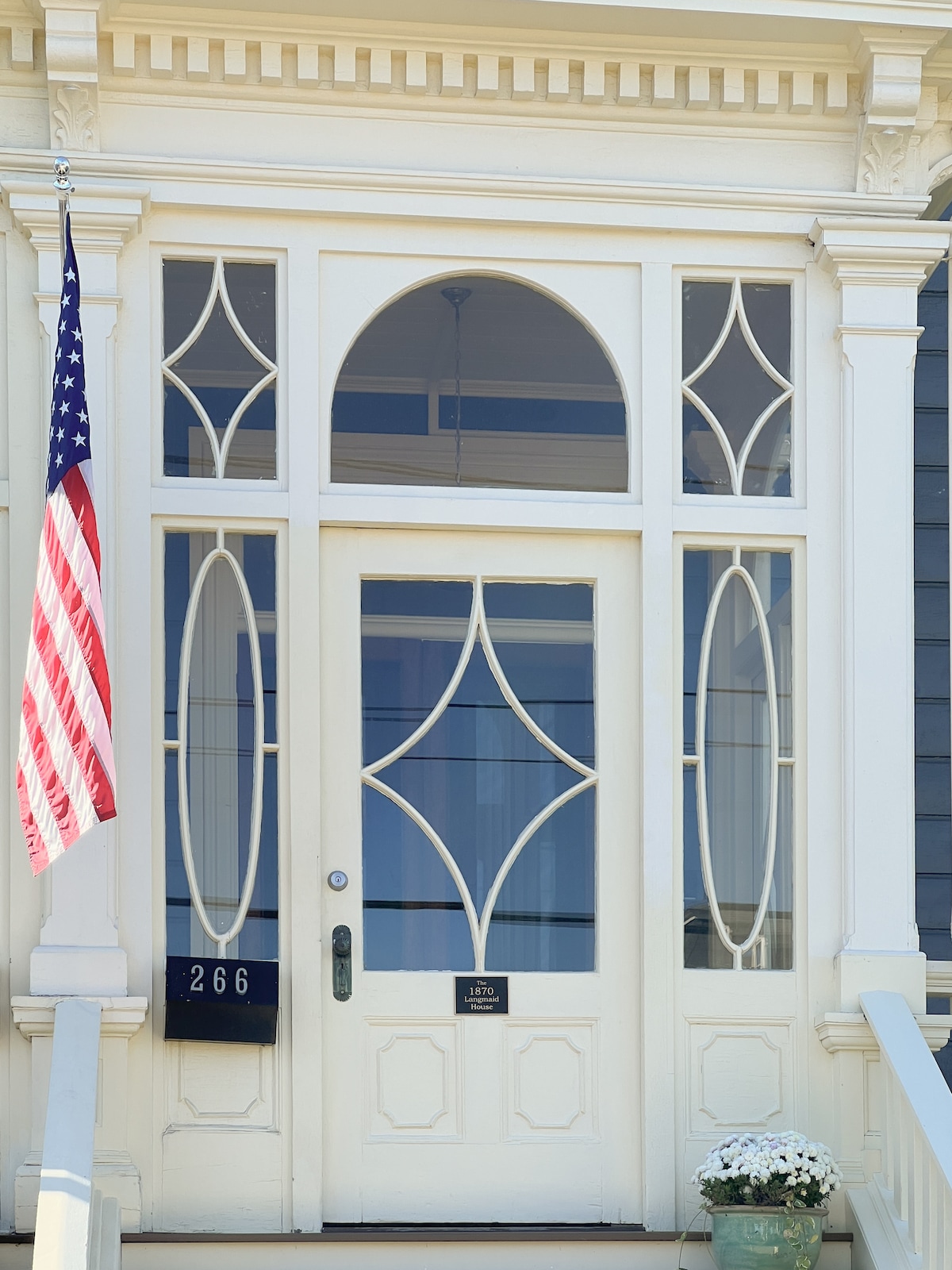 The main entrance features a large, ornate door with intricate glass designs that allow natural light to brighten the foyer. An American flag is displayed beside the door, and a small planter with white flowers sits at the base of the steps.