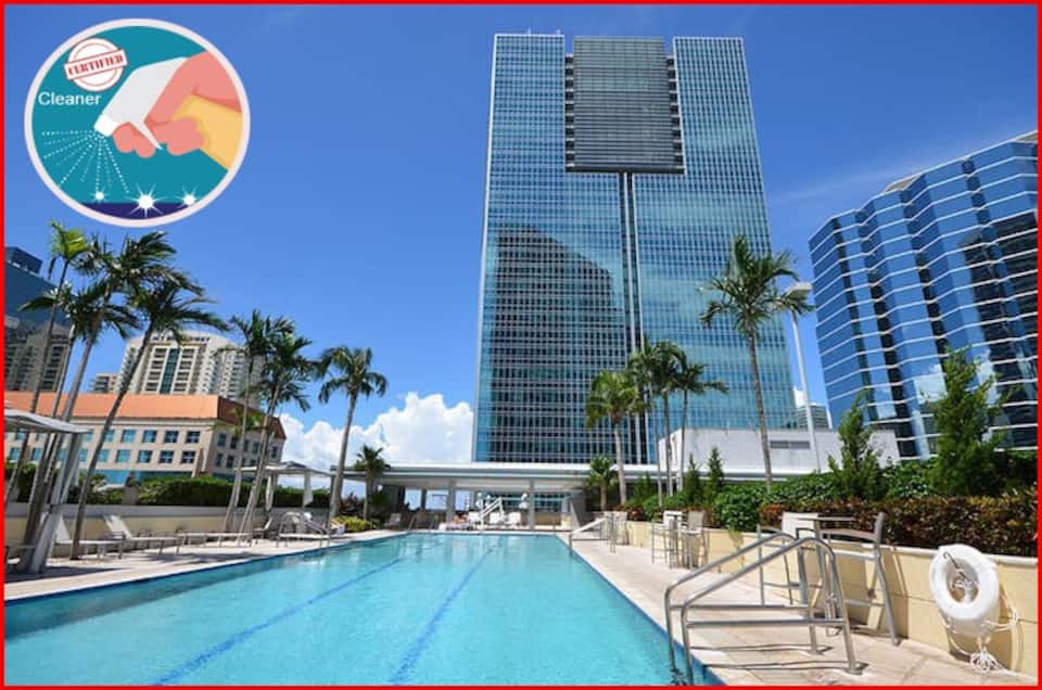 A large outdoor pool is situated amidst palm trees and modern high-rise buildings. The pool deck features lounge chairs and shaded seating areas, creating a relaxed atmosphere for guests. The clear blue sky adds to the inviting setting.