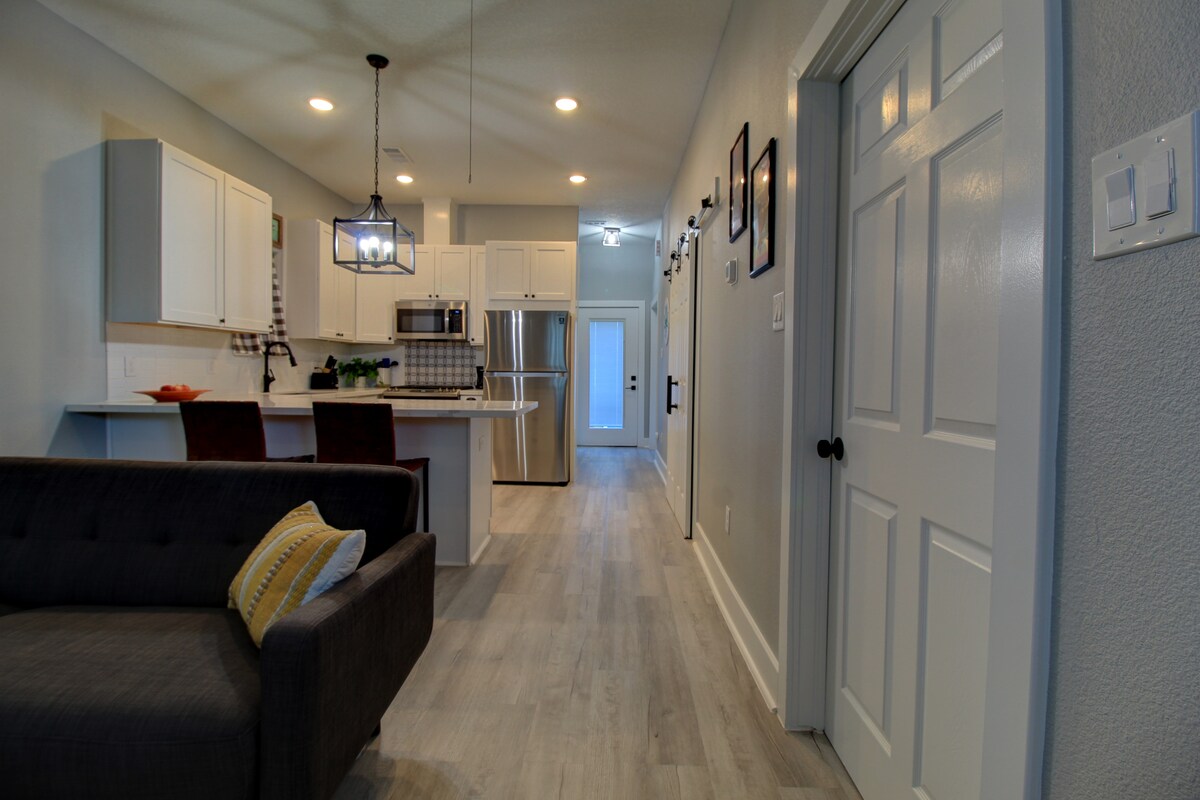 An open-concept living space is visible, featuring a cozy couch with a decorative pillow. A kitchen with modern appliances and a dining area with bar seating is highlighted in the background. Light-colored walls and warm lighting contribute to a bright and welcoming atmosphere.