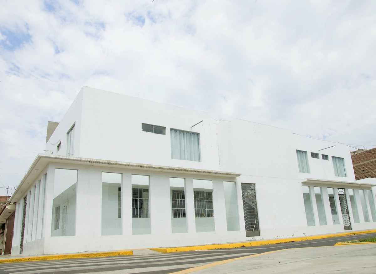 A contemporary building with a white exterior is seen from the street. Large windows are present, allowing for ample natural light. The structure is situated on a corner, with a wide sidewalk and a smooth roadway, contributing to an open and inviting ambiance.
