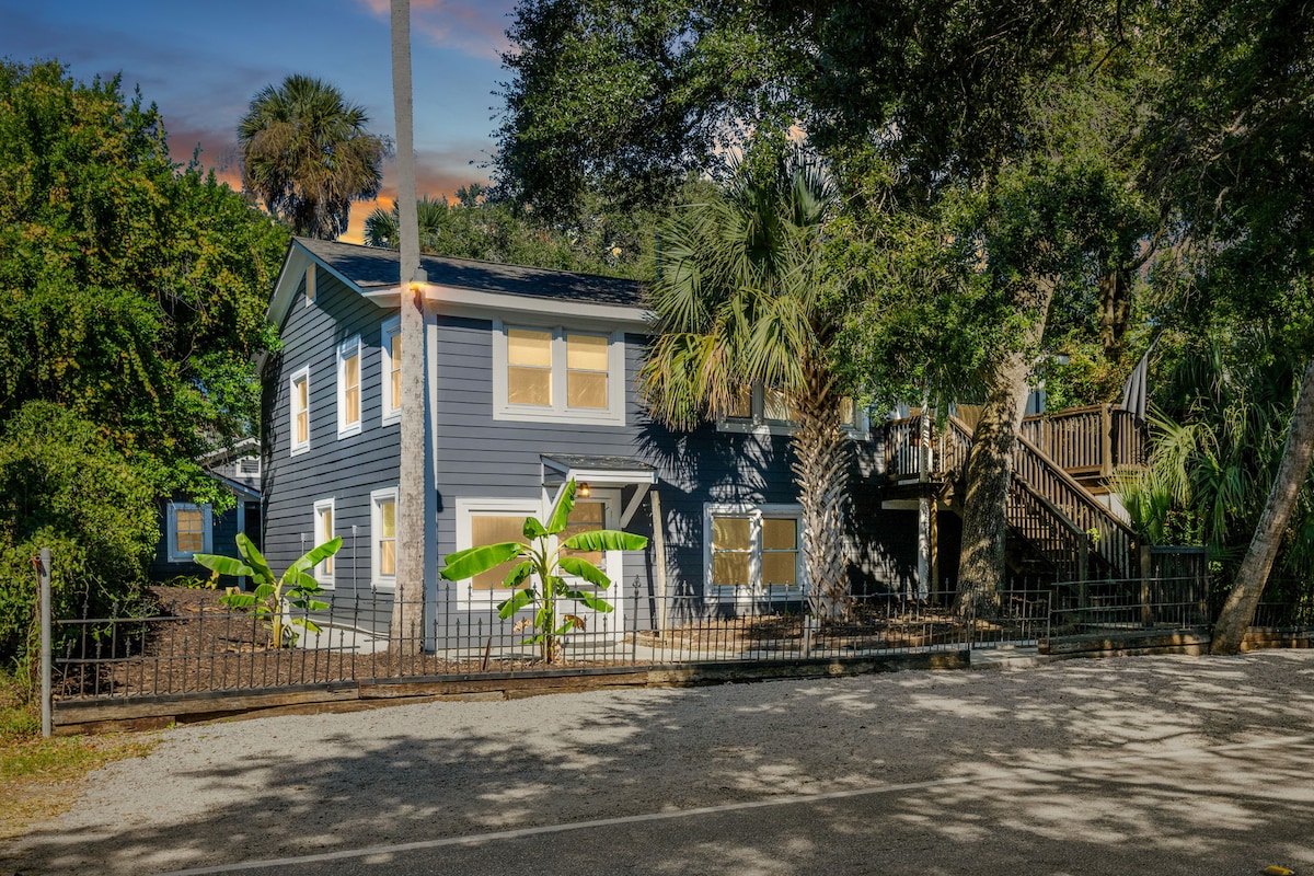 A two-story blue house is framed by lush greenery, showcasing palm trees and banana plants at the front. The entrance features a set of wooden stairs leading to the upper level, which is adorned with multiple large windows that allow ample natural light.