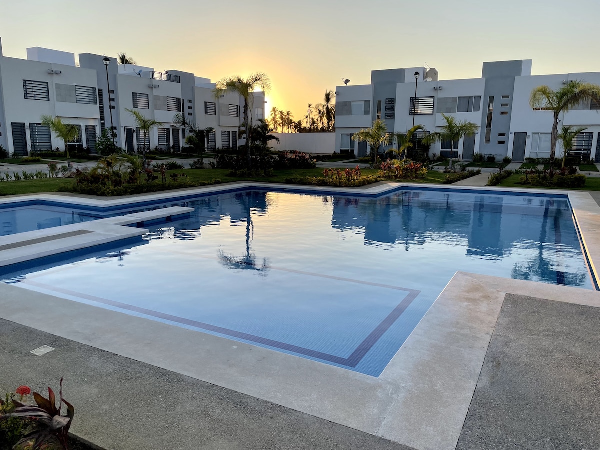 A large swimming pool is shown, reflecting the sunrise. Surrounding the pool are well-maintained green lawns and colorful landscaping. The modern buildings of the condominium are visible in the background, creating a serene atmosphere.