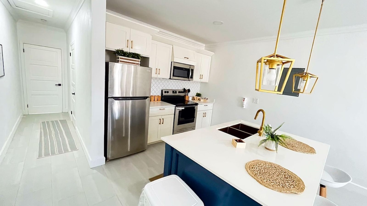 A modern kitchen is depicted, featuring stainless steel appliances, a spacious island with bar seating, and elegant pendant lighting. White cabinetry complements the light-colored tile floors. Decorative elements, such as natural fiber placemats, add a subtle touch to the contemporary design.