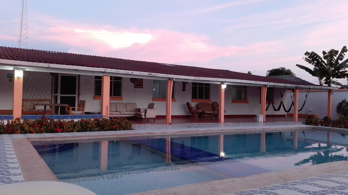 A serene swimming pool reflects the soft hues of the sunset. Surrounding the pool, comfortable seating areas provide relaxation options. The house features a covered porch with light-colored furnishings, framed by flowering plants and greenery, enhancing the tranquil atmosphere of the outdoor space.