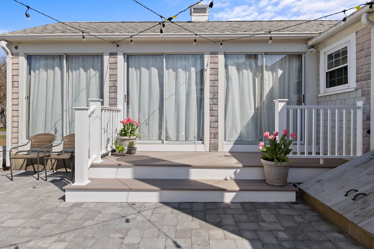 A spacious outdoor deck is shown, featuring sliding glass doors that lead to an interior space. Two potted plants with pink tulips are placed at the base of the steps. String lights are draped overhead, enhancing the inviting atmosphere.