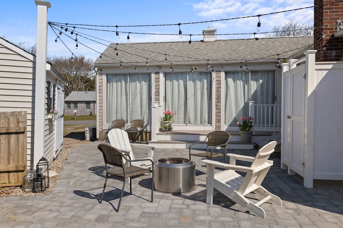 A paver patio is surrounded by seating arrangements that include four chairs and a fire pit at the center. The area is accented by hanging string lights and flower pots, with large glass windows in the background letting in natural light.
