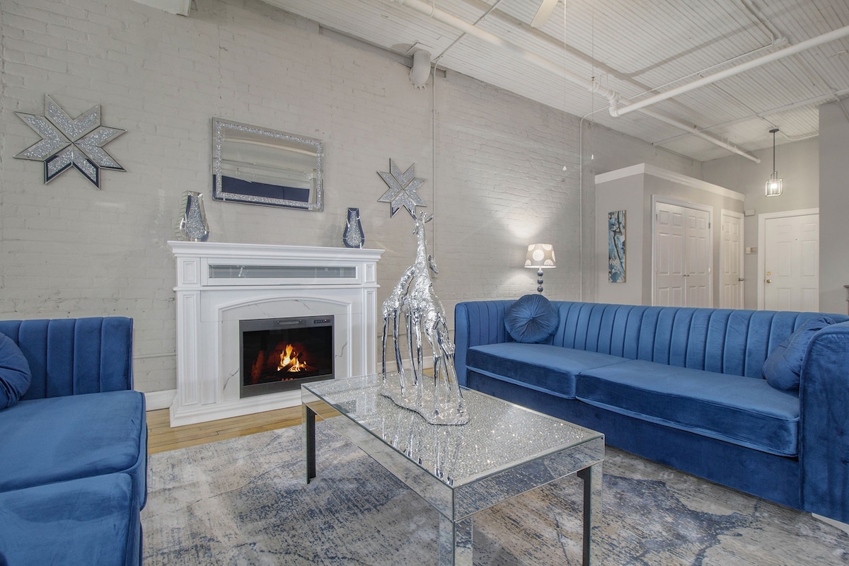 The living area features two deep blue velvet sofas facing a mirrored coffee table, which sits in front of a decorative fireplace. Soft lighting is provided by a table lamp with a silver base, and a large mirror reflects the room's design elements.