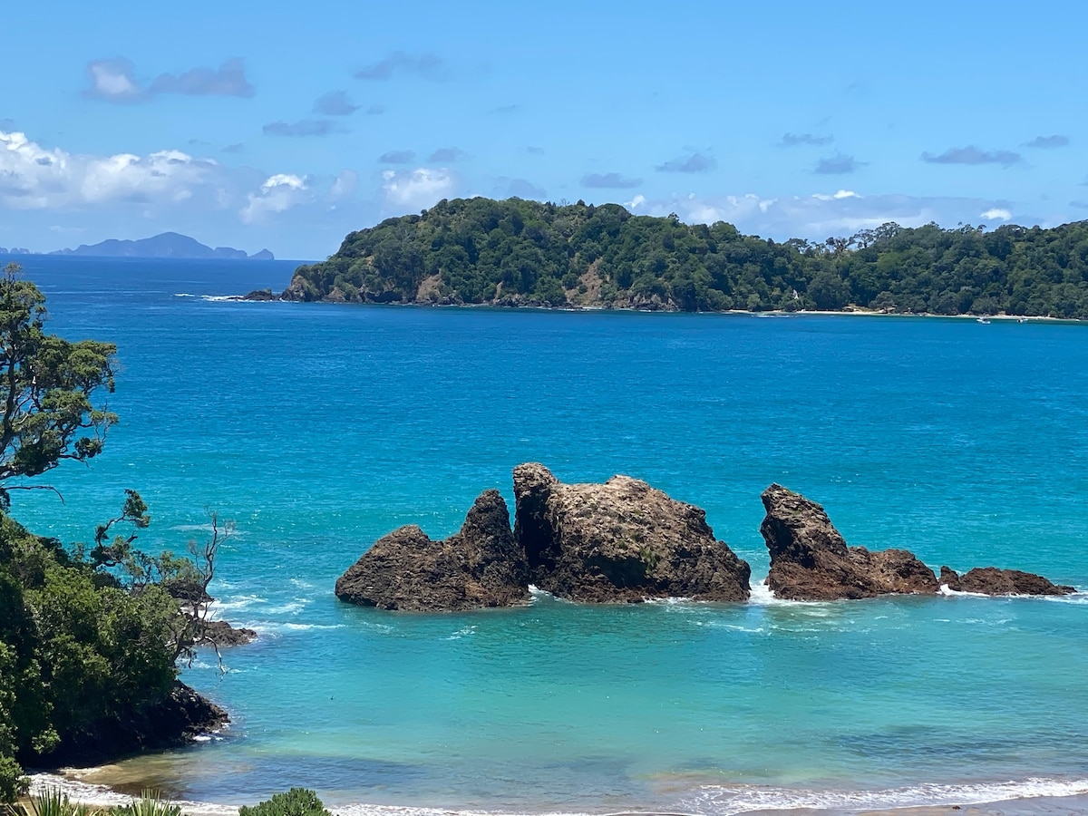 A panoramic view of the tranquil sea, showcasing a rocky outcrop rising from azure waters. Lush greenery outlines the shoreline in the distance, while the vibrant blue sky features scattered clouds. The scene captures a serene coastal landscape, inviting relaxation and connection with nature.