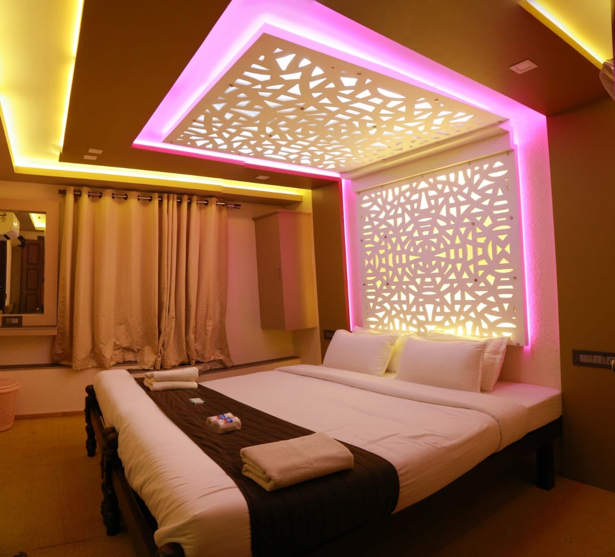 A brightly lit bedroom features a spacious bed with crisp white linens. Soft pink and yellow LED lights accentuate the modern ceiling design. Elegant curtains frame the window, while decorative wall patterns add a touch of artistry to the ambiance.