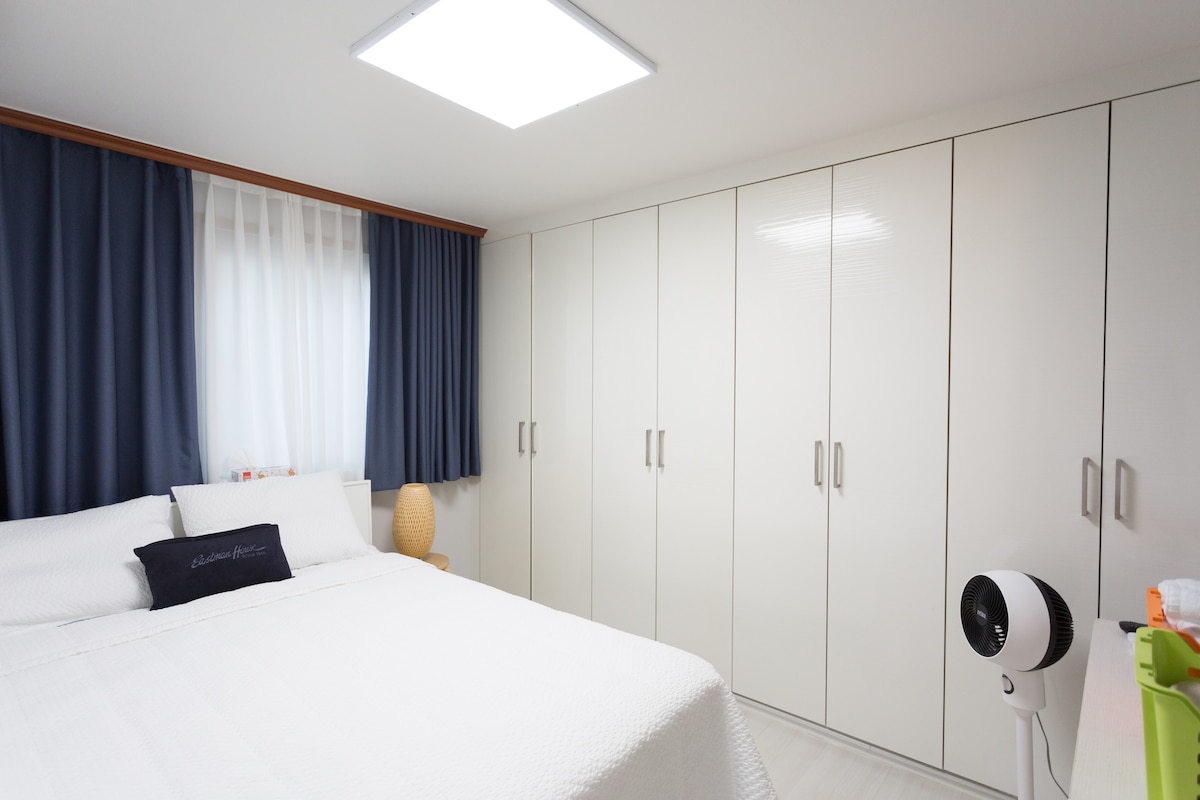A bright bedroom features a spacious bed with a white quilted coverlet and decorative pillows. A large window covered with sheer curtains allows natural light to enter. White wardrobes with multiple doors line one wall, while a round lamp sits beside the bed.