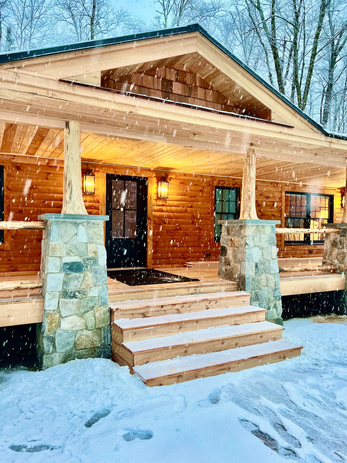 The inviting entrance of the cabin showcases wooden steps leading up to a covered porch, framed by natural stone pillars. Soft snowflakes fall gently amidst the serene setting, while warm light glows from the windows, highlighting the cabin's rustic charm against the snowy backdrop.