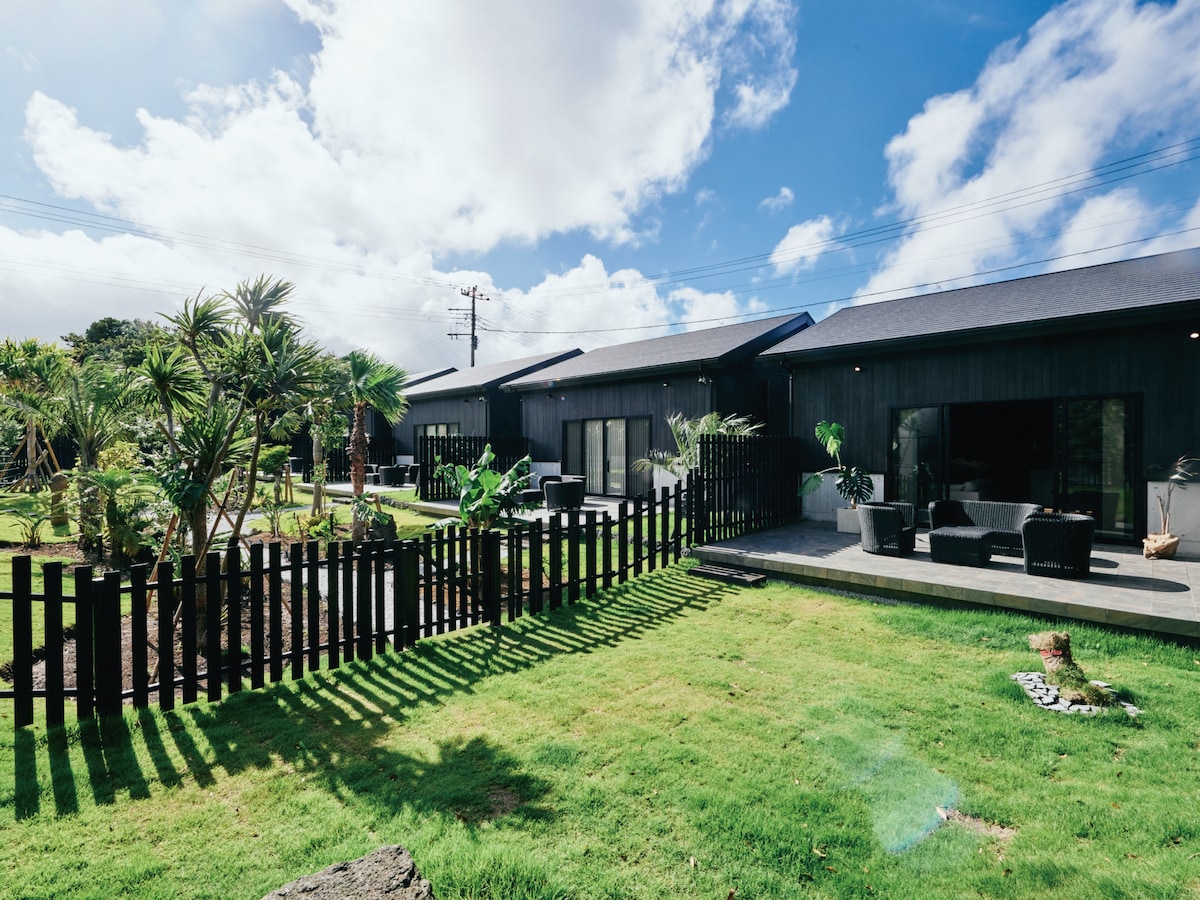 A spacious outdoor area features a neatly manicured lawn surrounded by tropical plants. Modern seating arrangements are positioned on a patio, offering a relaxing atmosphere. A black wooden fence outlines the space, contributing to the tranquil setting under a bright blue sky.