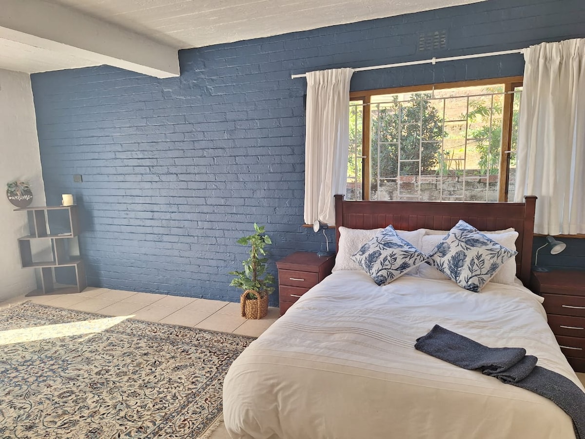 A spacious bedroom features a large bed with decorative blue-themed pillows. Natural light pours in through a window, illuminating the textured blue wall. A small plant in a woven basket and a neatly arranged rug enhance the room's inviting atmosphere.