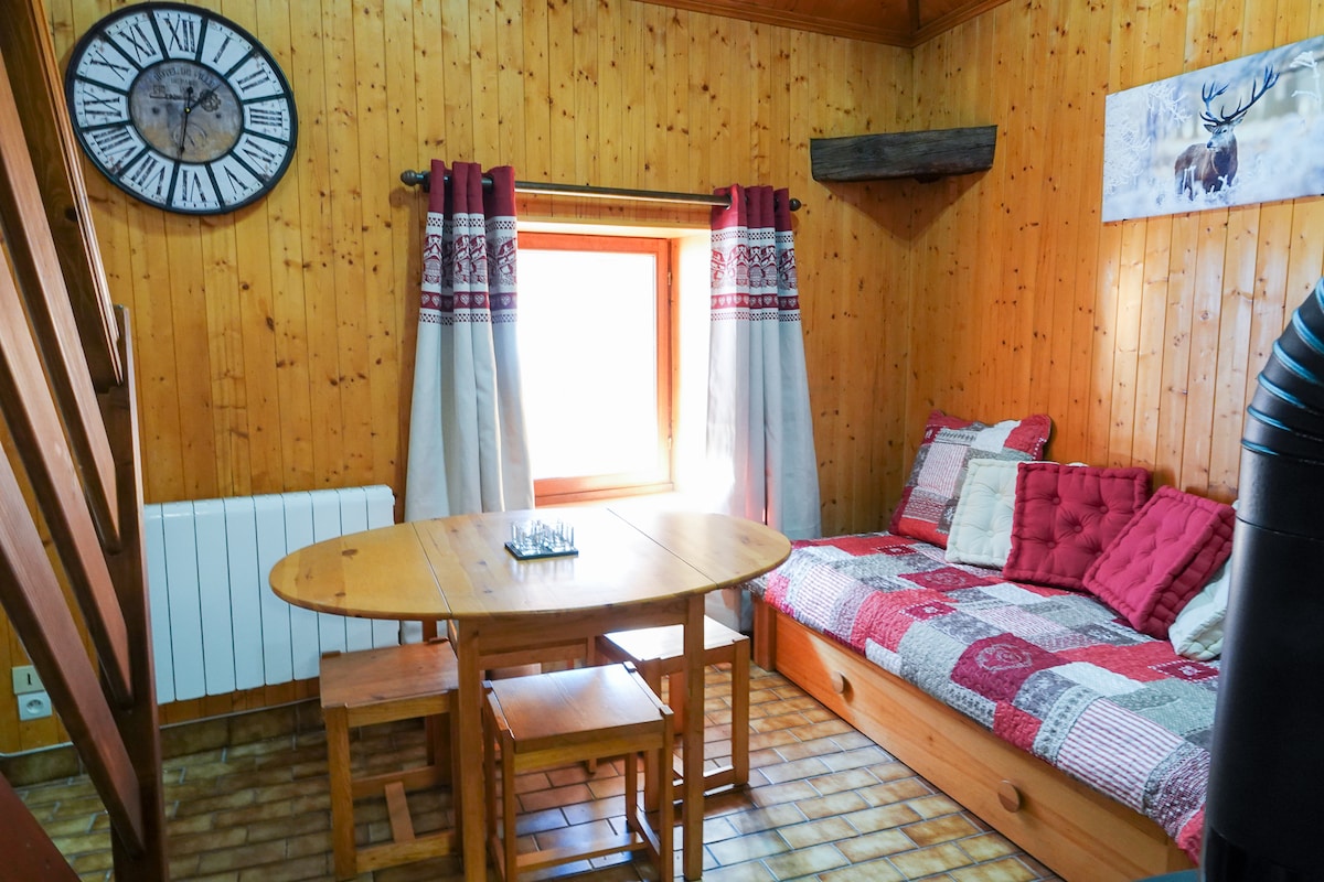 A wooden interior is captured, showcasing warm tones and a cozy seating area featuring a seating arrangement with cushions. A circular table is positioned nearby, illuminated by natural light entering through a window adorned with patterned curtains. A clock decorates the wall, enhancing the rustic ambiance.