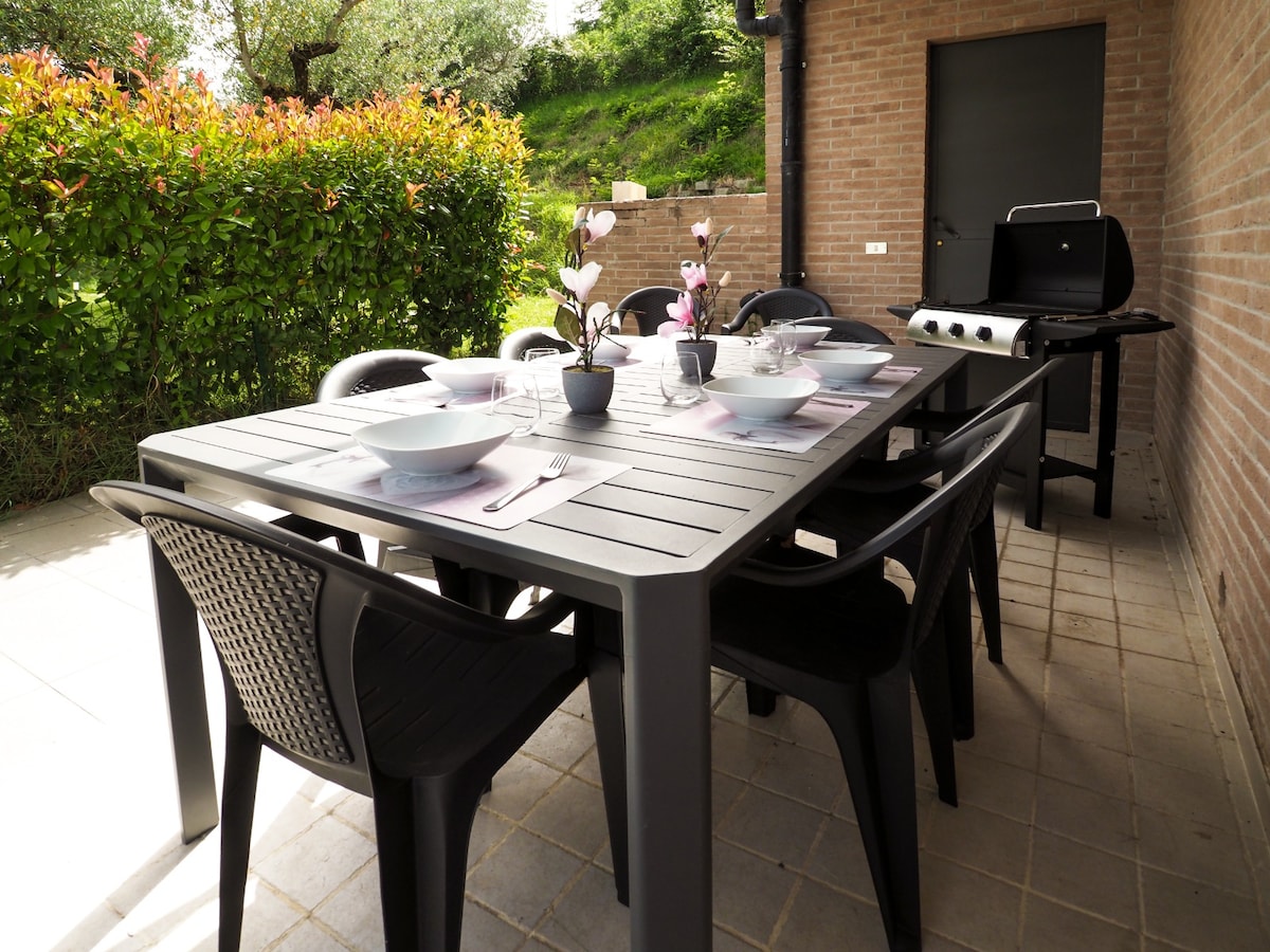 An outdoor dining area is presented with a large grey table set for six, featuring white plates and glassware. The background displays lush greenery and a barbecue grill, providing an inviting space for al fresco meals.