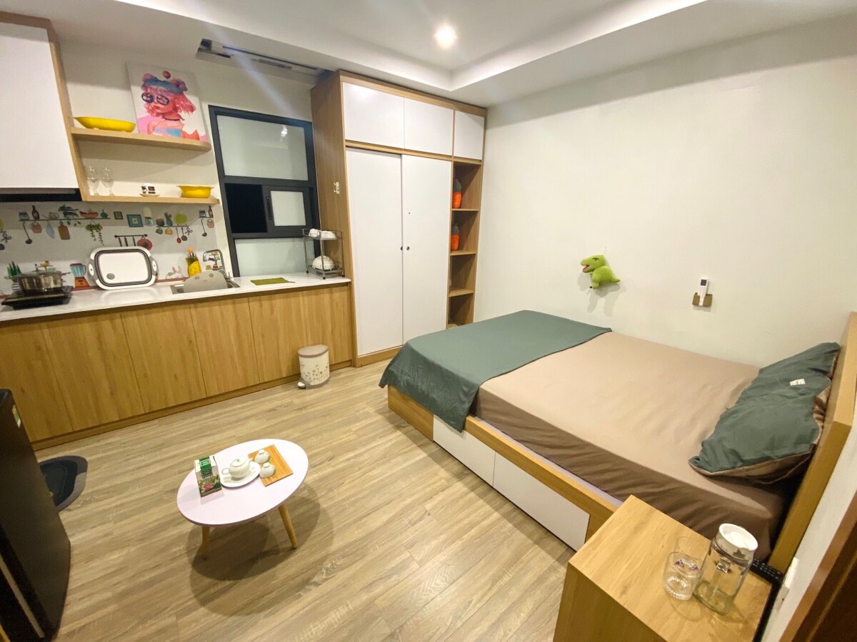 A well-organized studio apartment features a spacious bed with a muted blanket. A small coffee table is positioned in front of the bed, accompanied by two glasses. A compact kitchenette with essential utensils is visible, along with a wardrobe that provides additional storage.