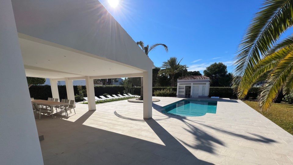 A spacious outdoor area features a clear swimming pool surrounded by sun loungers. The sun casts soft shadows on the patio, while palm trees and a well-maintained lawn enhance the serene setting. A covered dining space is also visible, offering ample seating.