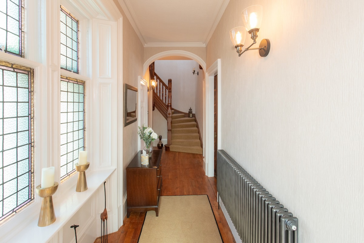A welcoming entryway features elegant stained glass windows, allowing natural light to filter in. A wooden console table is positioned against one wall, complemented by decorative candles. The hallway leads towards a staircase with a wooden handrail, enhancing the inviting atmosphere of the space.