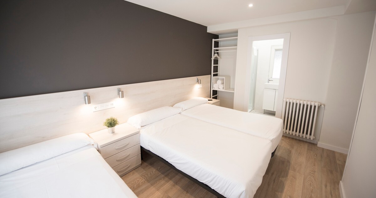 A bright room is presented with three single beds arranged neatly against a dark accent wall. Each bed is paired with a bedside table featuring soft lighting. A closet area is visible on the side, and a bathroom door leads to a private bathroom.