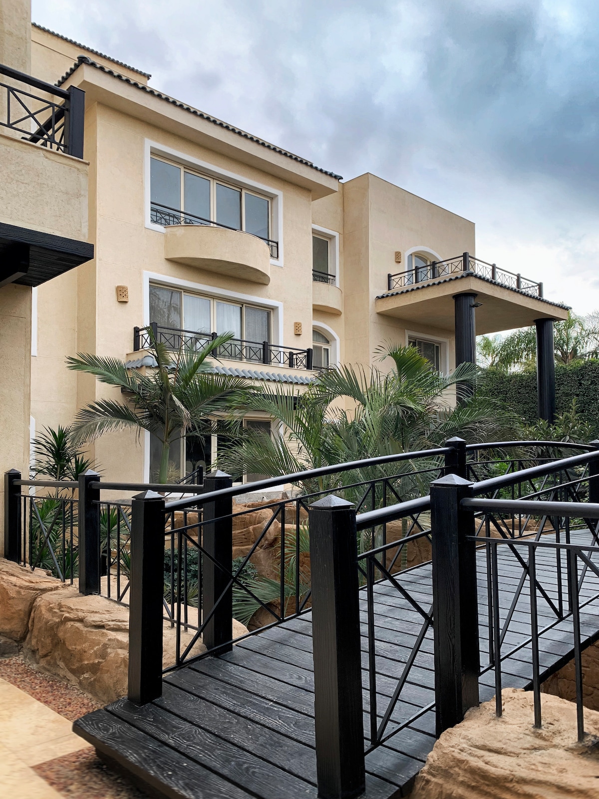 The exterior of the villa is showcased, featuring a multi-story structure with large glass balconies and palm trees. A wooden bridge leads to the entrance, enhancing access to the garden area surrounding the home.